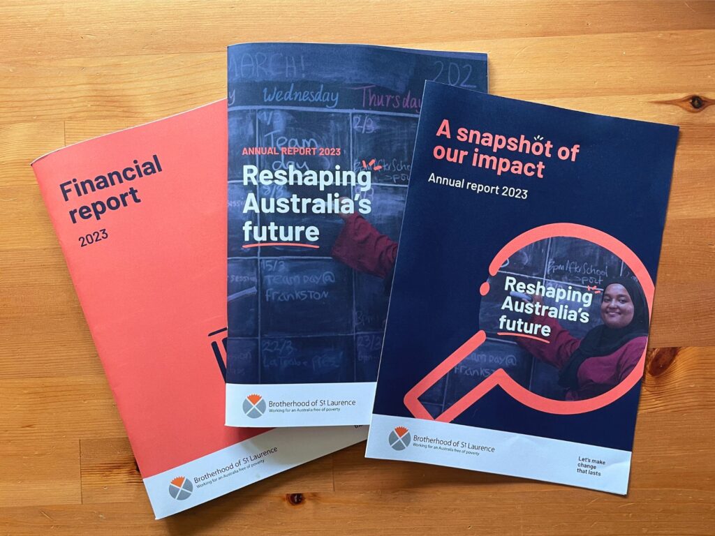Real photograph of three Annual reports from Brotherhood of St. Laurence (BSL) fanned out on a wooden table, including the Financial Report 2023, 'A snapshot of our impact' summary report, and 'Reshaping Australia's Future' full version print report.